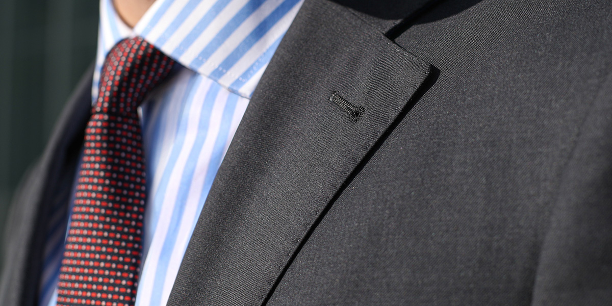 Particolare sul bavero di una giacca, indossata su camicia a righe blu e bianche e una cravatta micropuntinata.