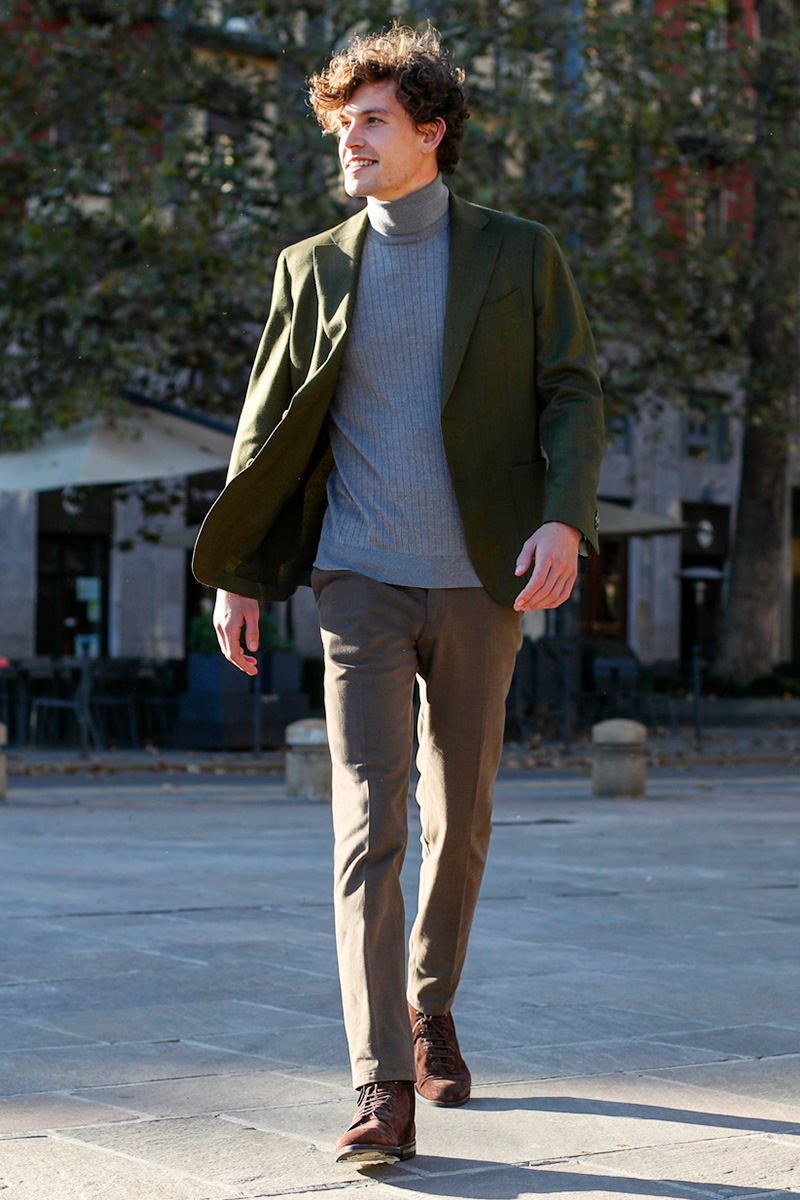 A man wears a green blazer and bespoke brown trousers with a light blue ribbed turtleneck sweater