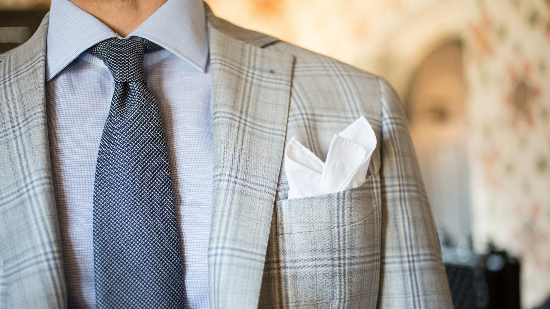 Dettaglio su una giacca da uomo Principe di galles, camicia azzurra, cravatta blu e pochette bianca piegata a sbuffo