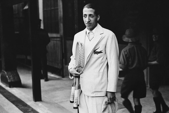 Renè Lacoste wears a white suit over a polo shirt with, wielding two tennis rackets