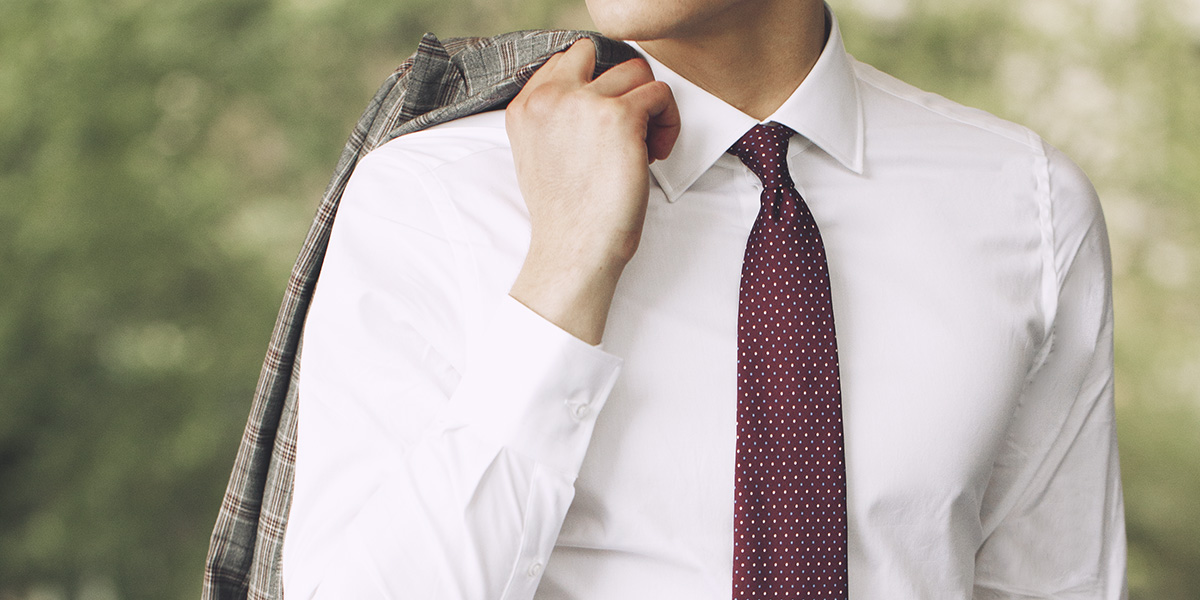 Dettaglio outfit uomo: camicia bianca, cravatta rossa puntinata e giacca grigia in principe di galles sulla spalla.