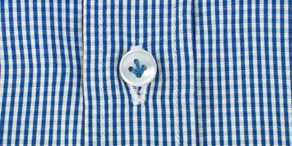 Dettaglio sul bottone di una camicia a righe blu e bianche cucito con filo azzurro. 
