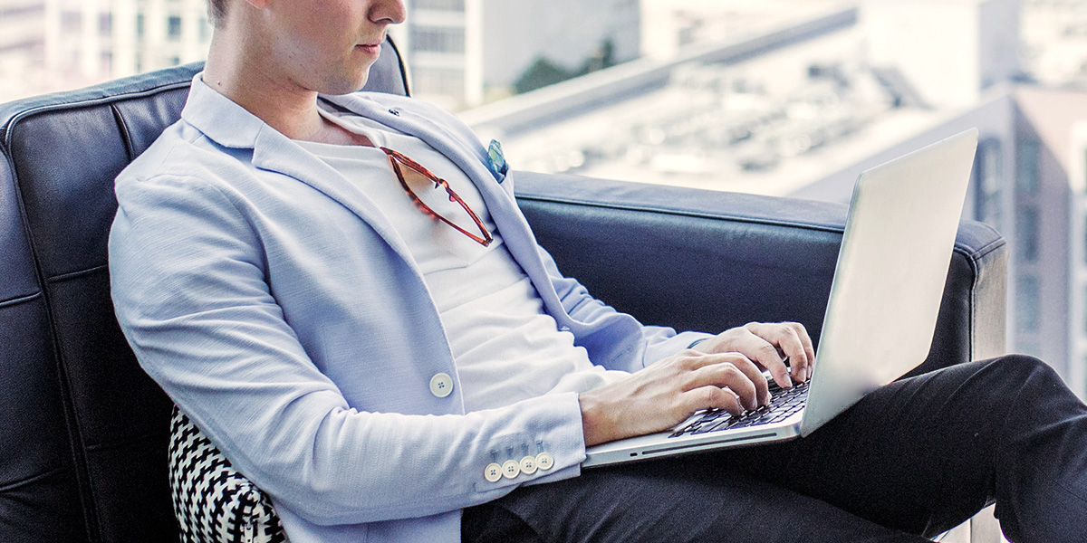 Uomo seduto su una poltrona indossa una t-shirt bianca e un blazer azzurro  mentre lavora d un computer portatile