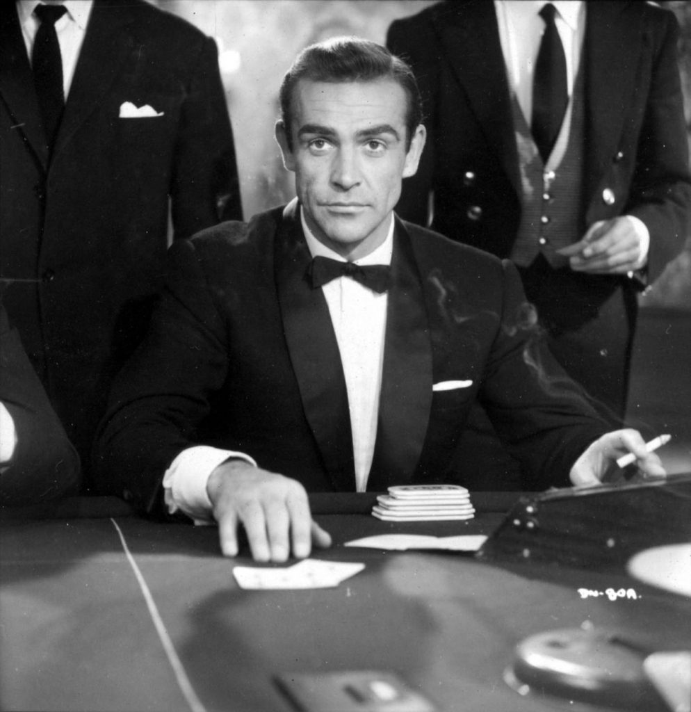 Actor Sean Connery plays James Bond 007 in a scene from the movie Casino Royale (1967) wearing a black tuxedo, a white flat-fold pocket square, while smoking a cigarette sitting at the game table
