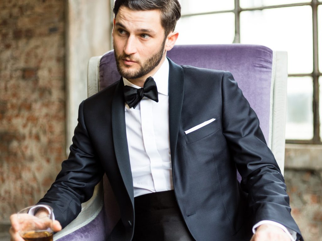 Man sitting on a purple armchair, wearing a black tuxedo by day with white shirt and black bow tie