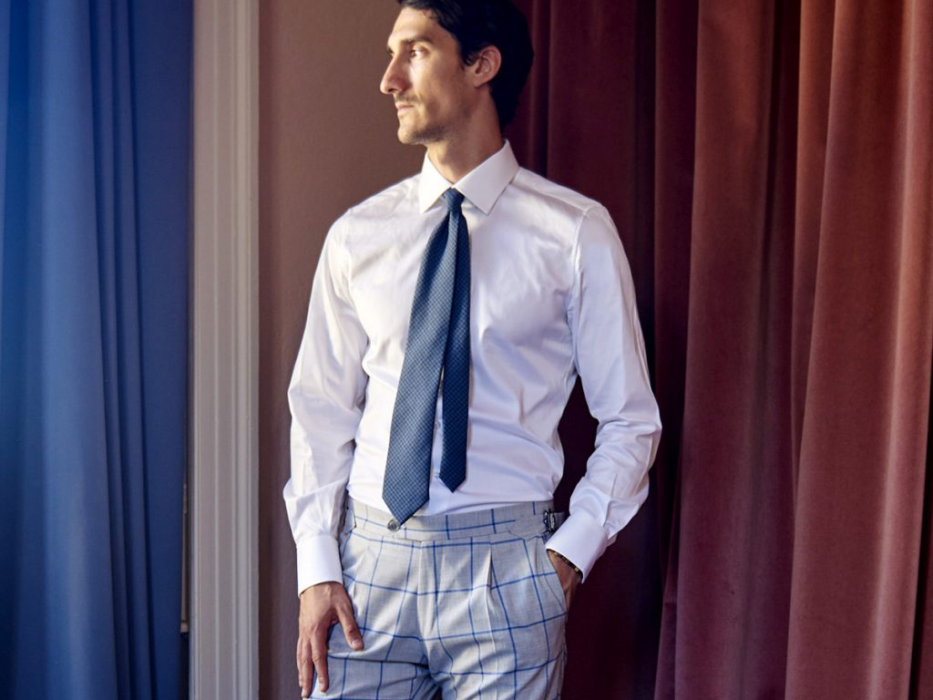 Un uomo indossa una camicia bianca, cravatta blu senza giacca e pantaloni a quadri.