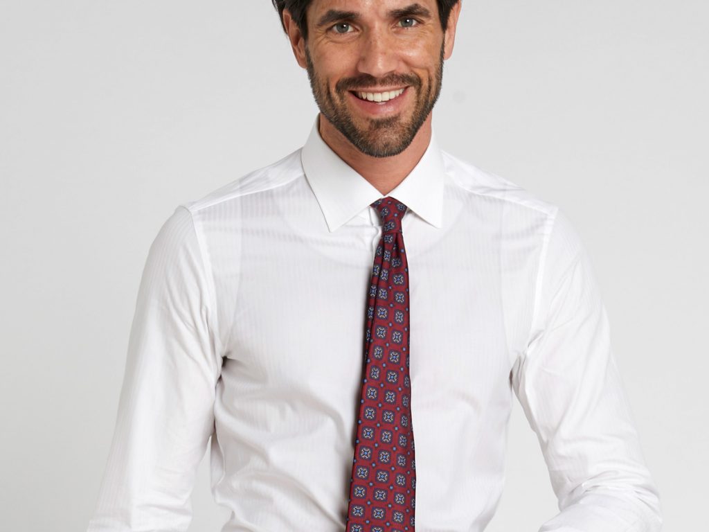 A man wears a white shirt with a transparent tank top and burgundy patterned tie