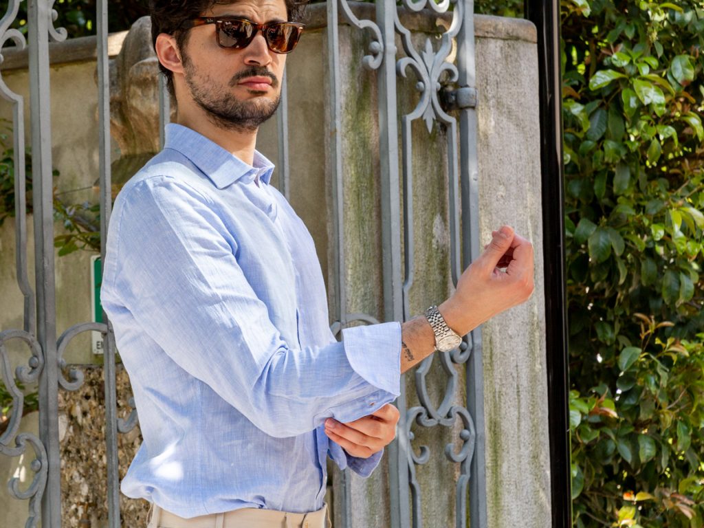 A man in sunglasses about to roll up the sleeves of his blue shirt